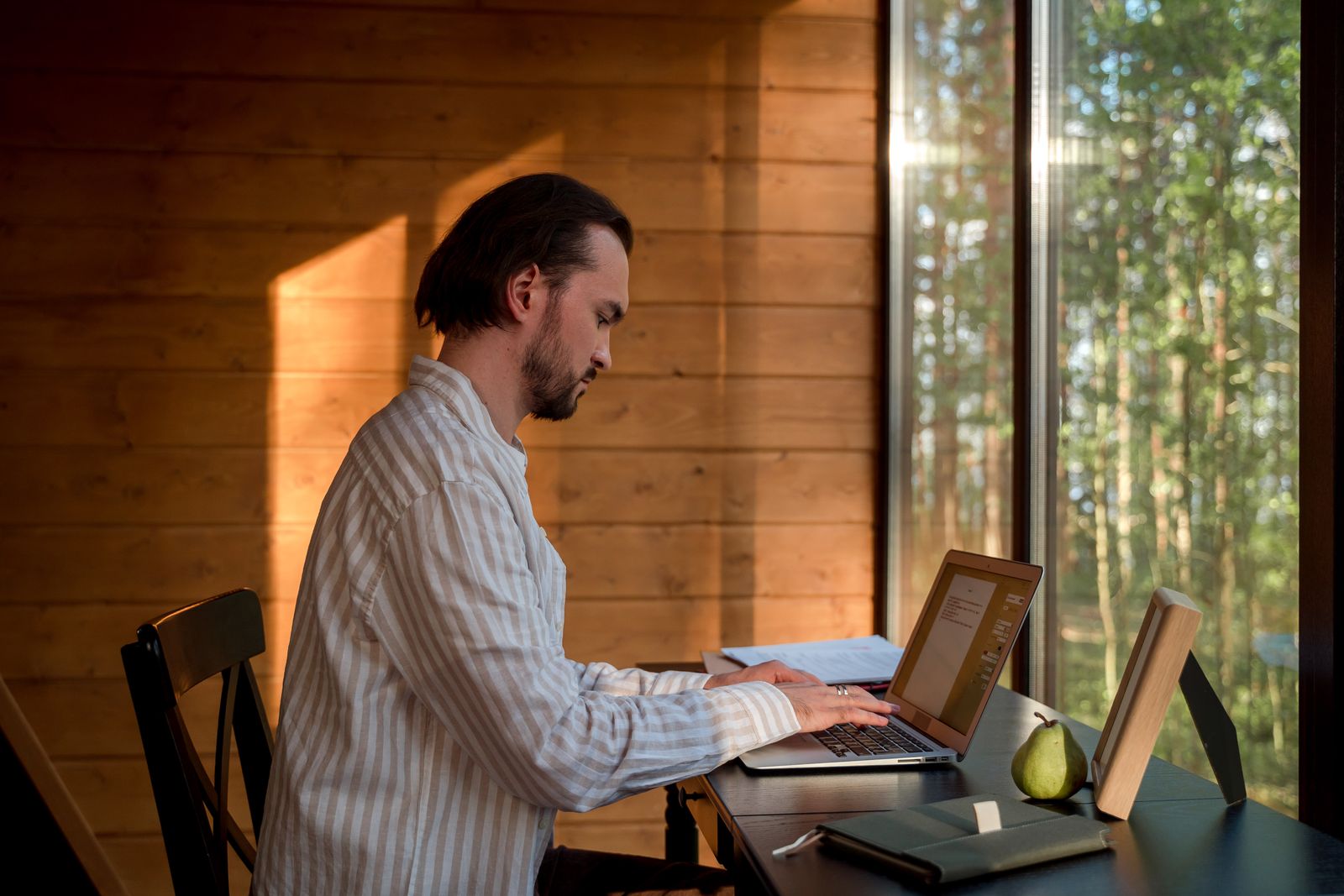 A man typing on a laptop at a home desk by a window, in soft natural light
