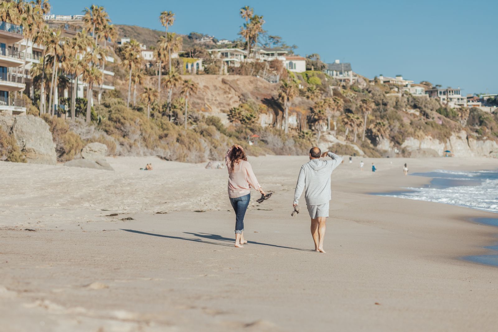 A couple walking along a sunny beach below a hillside of homes